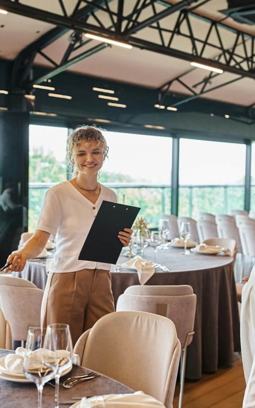 event organizer with clipboard pointing at table with festive setting near woman in banquet hall