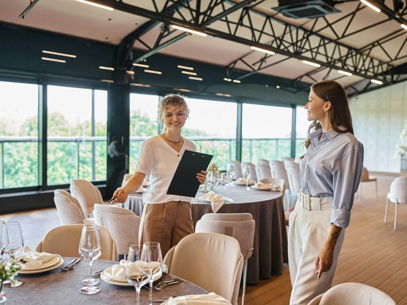 event organizer with clipboard pointing at table with festive setting near woman in banquet hall